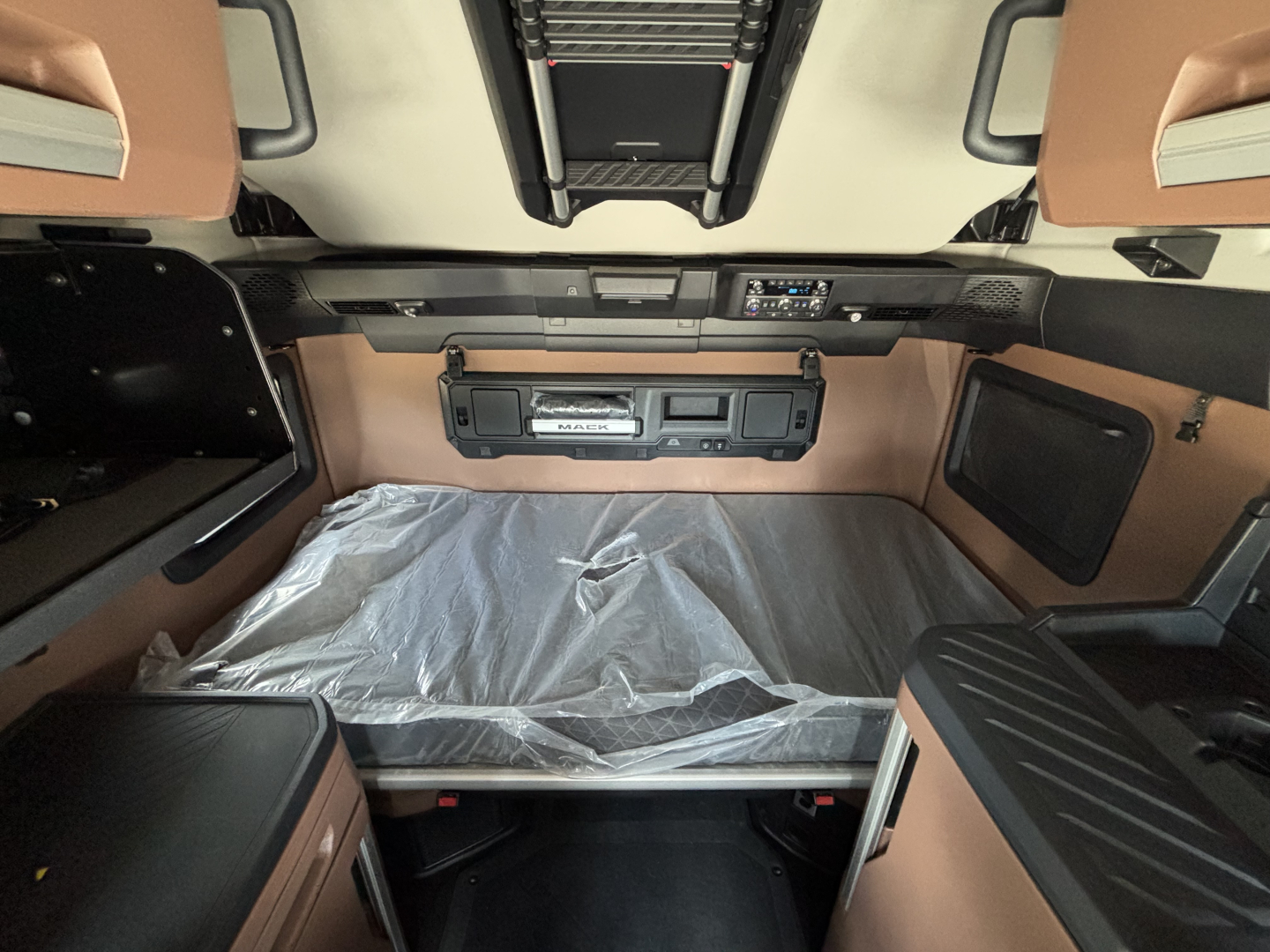 Cab interior of a new Mack PR64T76H truck sleeper showing overhead controls and storage compartments with brown upholstery and plastic-covered bunk.