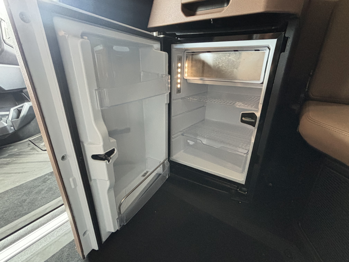 Trunk/cabinet area of a 2027 Mack PR64T76H showing a compact fridge/freezer unit inside a sleeper cab with open fridge door and empty shelves.