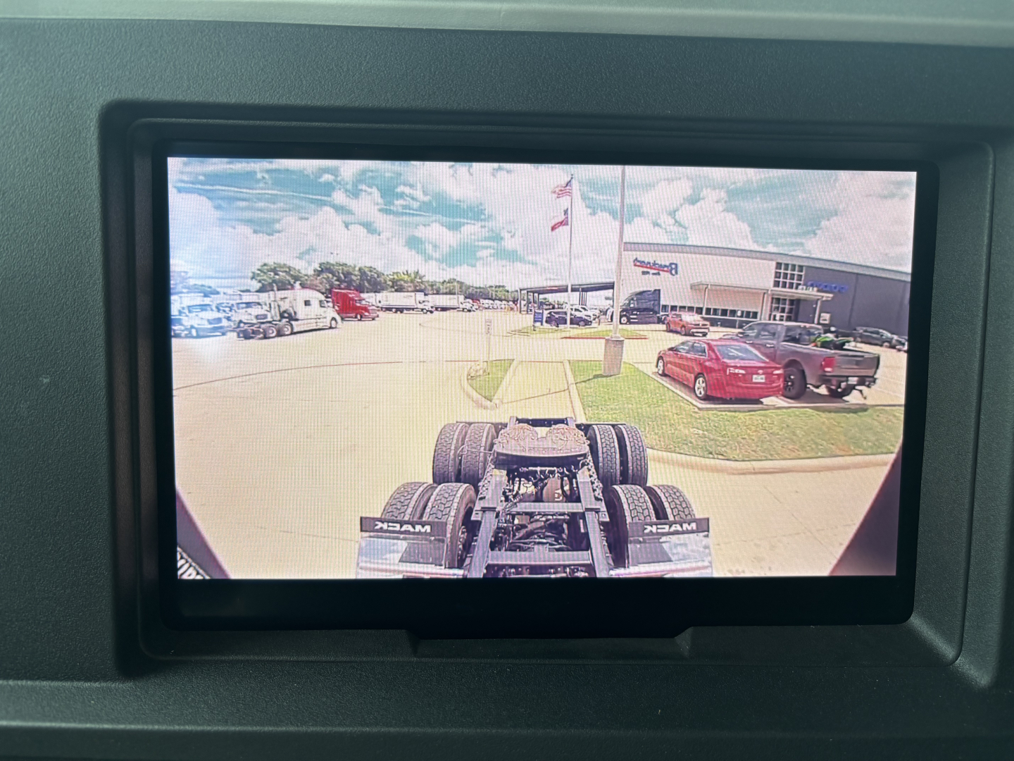 Truck cab rear view showing fifth wheel and rear dual tires in an outdoor lot; on-screen display from a monitor showing a dealership with cars and flags.
