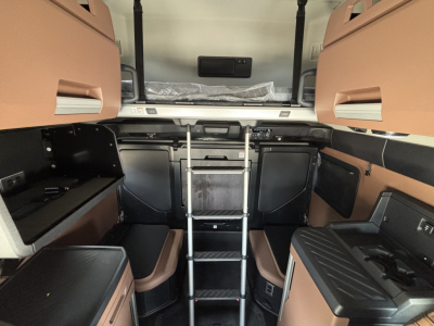 Truck cab interior view of a 2027 Mack PR64T76H sleeper, with a center ladder to the upper bunk, storage cabinets on both sides, and beige-brown upholstery.
