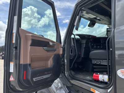 Truck cab interior with open door showing Mack PR64T76H dashboard and steering wheel, driver seat, and entry area.