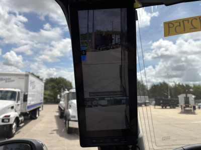 Truck cab interior showing large vertical display screen mounted in dashboard with reflection of yard and trucks outside.
