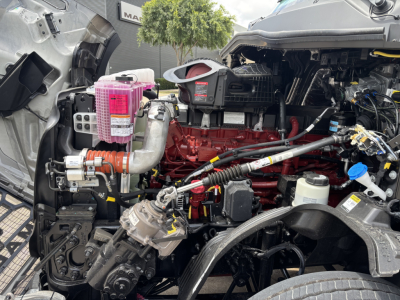 Mack PR64T76H engine bay showing pink coolant reservoir, exposed red engine block, hoses, and various components.
