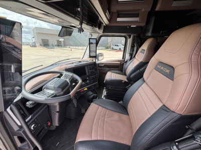 Truck cab interior showing steering wheel, dashboard, and two tan leather Mack seats with 'MACK' embroidery in a new Mack PR64T76H conventional sleeper.