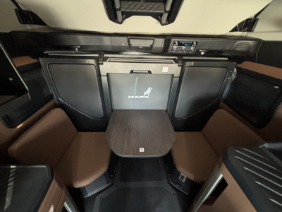 Mack PR64T76H sleeper cab interior showing dashboard, salt-brown seating, center table, and driver area with exposed storage slots.