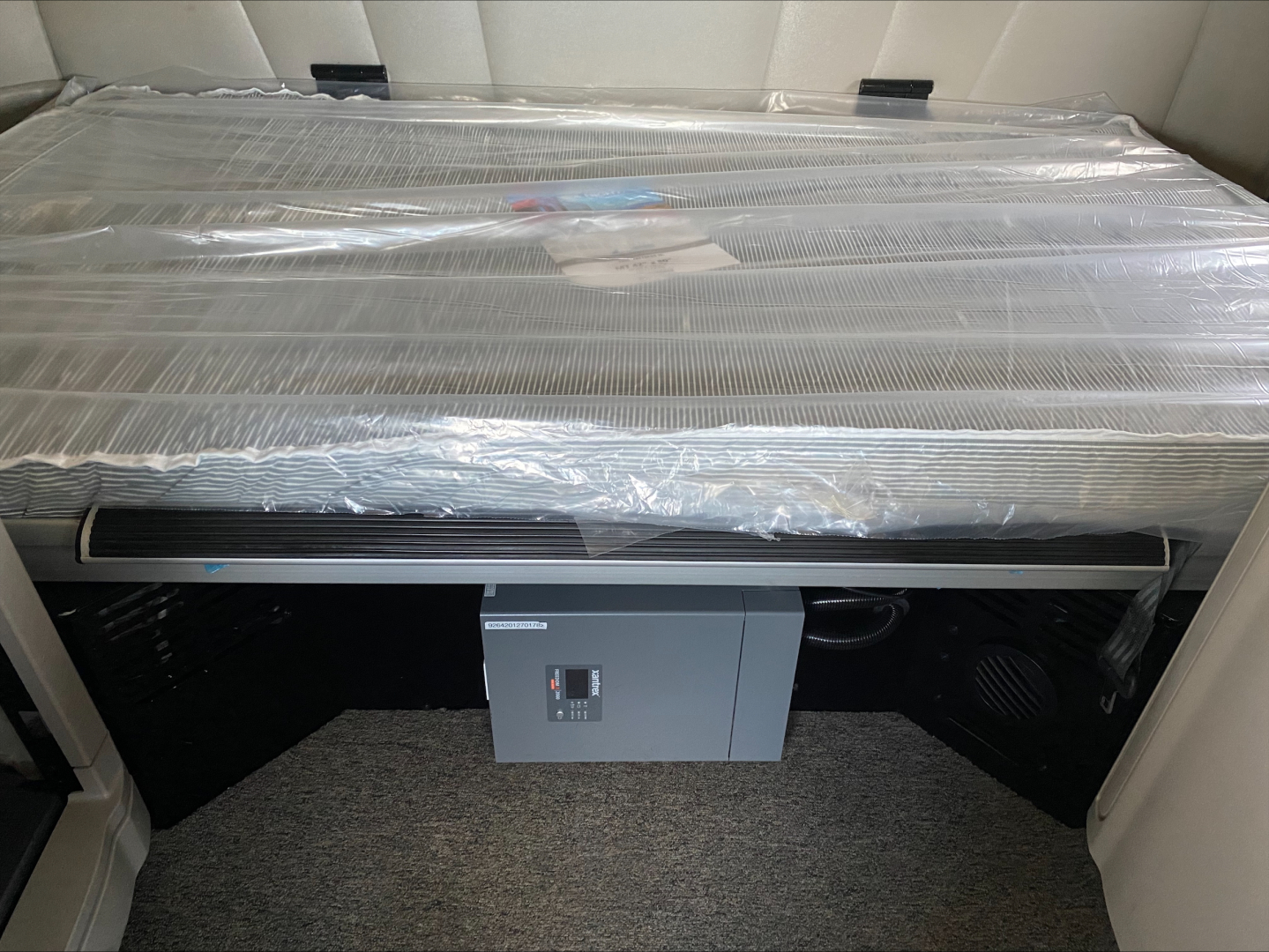 Conventional sleeper truck interior with a large radiator core and control box mounted below, wrapped in plastic; visible gray carpeting and beige seating behind.