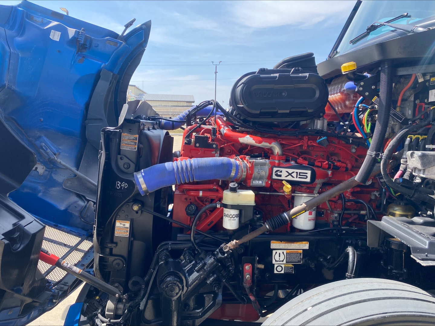 Peterbilt 579 engine bay showing Cummins X15 diesel engine with red block, blue coolant hose, air intake, and various reservoirs under hood.