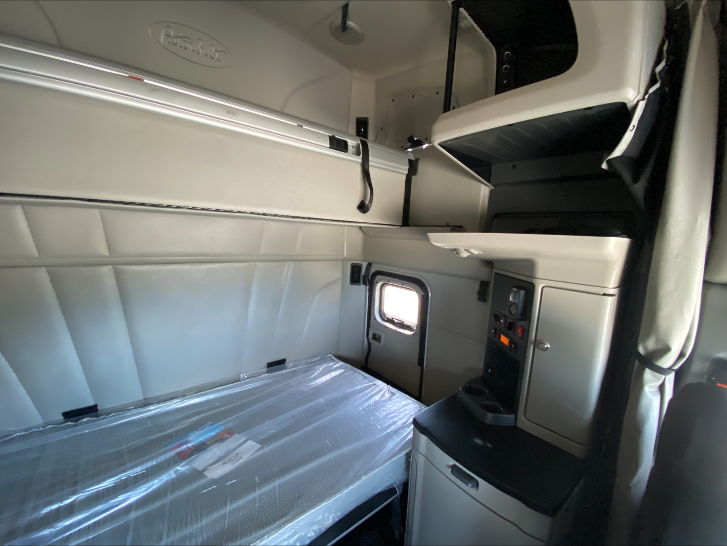 Peterbilt 579 sleeper cab interior showing bunk bed with plastic-wrapped mattress and upper storage shelf, side control panel, and small window.