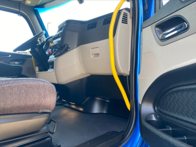 Truck interior cab view of a 2022 Peterbilt 579 with blue exterior, showing the driver seat, dashboard, steering wheel, and yellow door grab bar.