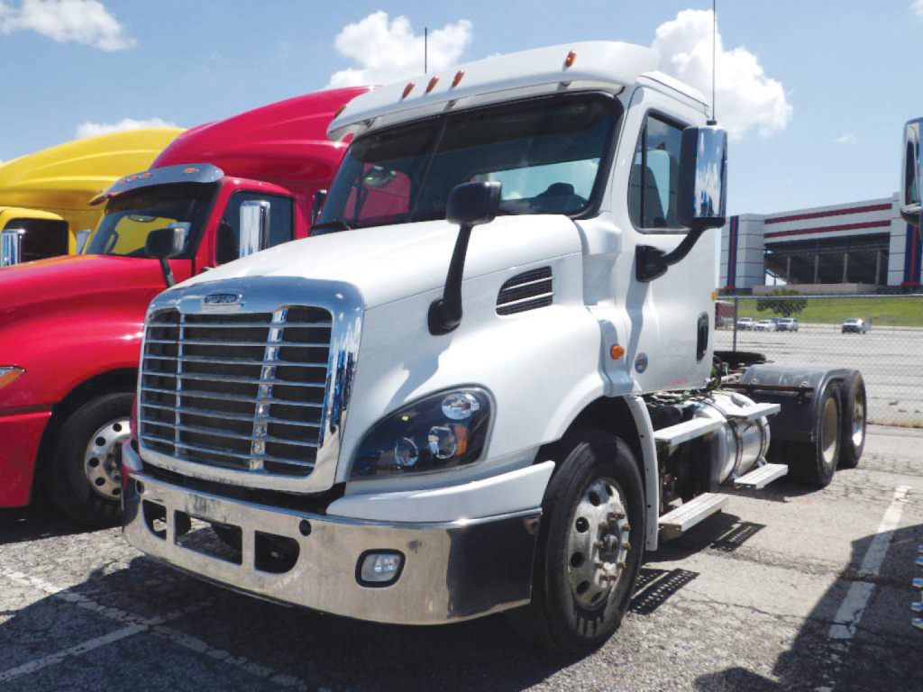 White Freightliner Cascadia Daycab truck in a dealership lot, front view showing chrome grille, bumper, and cab mirrors; other colorful trucks in background.
