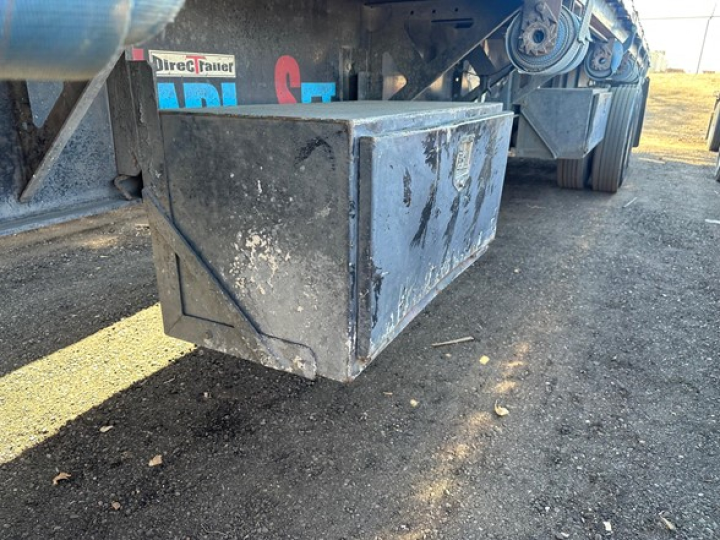 Flatbed trailer undercarriage showing a large rectangular toolbox mounted underneath and part of the wheel assembly in the background.