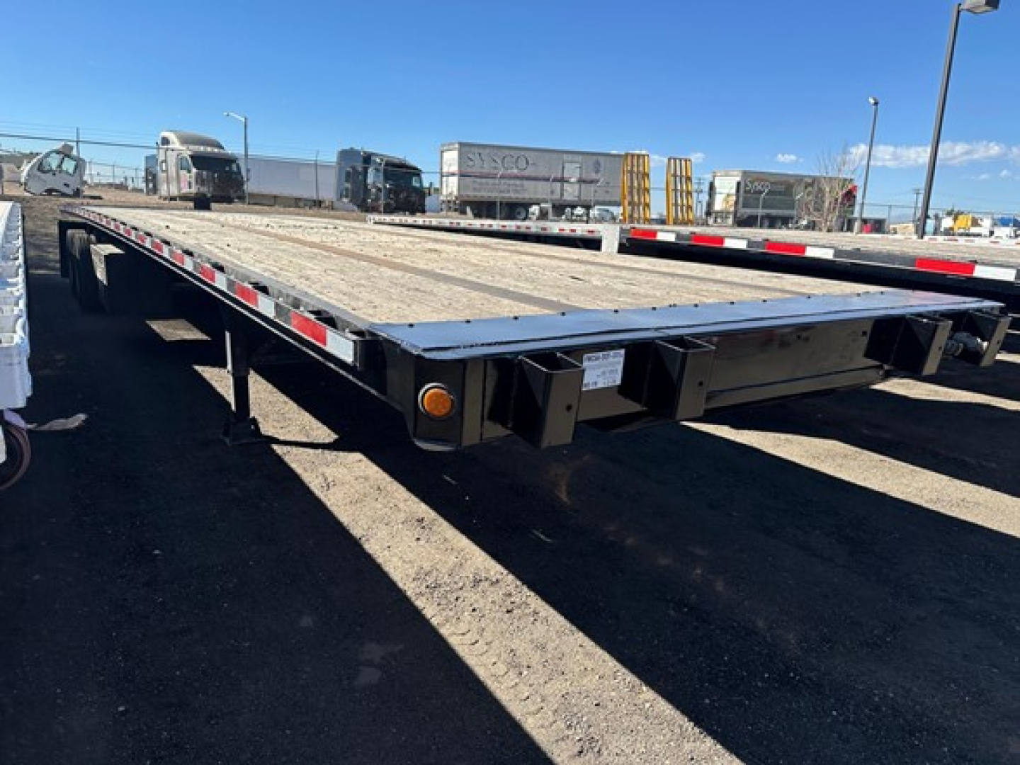 Direct Steel flatbed trailer in a yard, showing the long flat deck, rear bumper with lights, and side rails; multiple other trailers and trucks visible in the background.