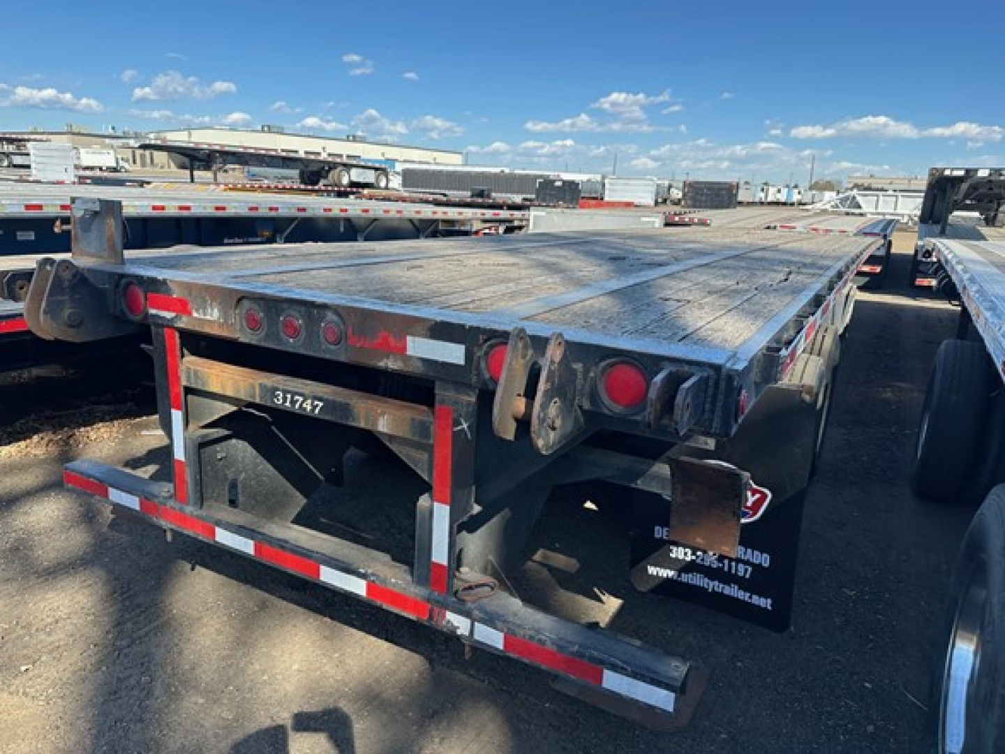 Flatbed trailer showing rear end with taillights, reflective tape, and dark metal deck; visible text on side panels includes contact graphics and numbers.