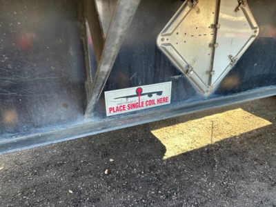 Trailer side showing sticker reading PLACE SINGLE COIL HERE and a diamond-shaped access panel on blue steel flatbed