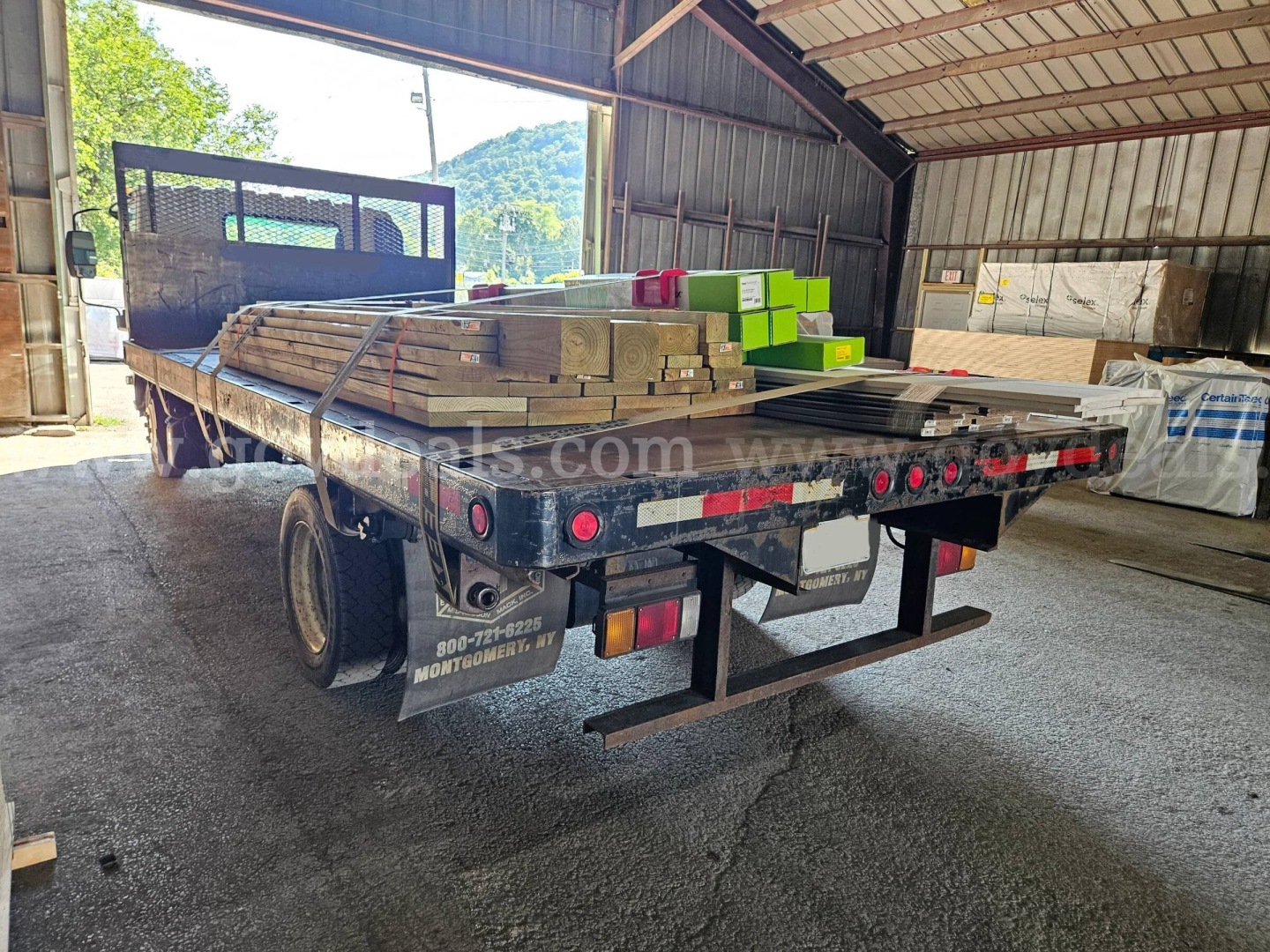 Isuzu NQR dump truck bed with lumber and materials loaded on flatbed, rear view inside a warehouse.
