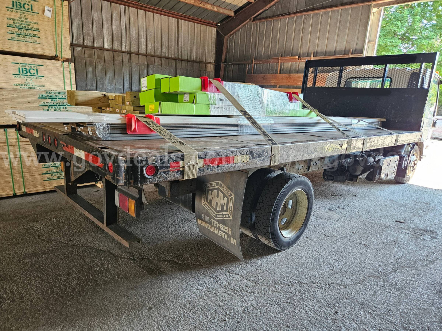 Isuzu NQR dump truck flatbed with cargo and stacked boxes in a storage shed; rear view showing dual rear wheels and flatbed with railing
