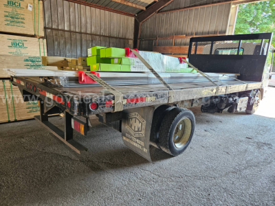 Isuzu NQR dump truck flatbed with cargo and stacked boxes in a storage shed; rear view showing dual rear wheels and flatbed with railing