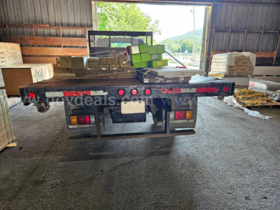 Isuzu NQR truck flatbed rear view with stacked boxes and pallets in a warehouse; rear lights and bumper visible; license plate area blank.