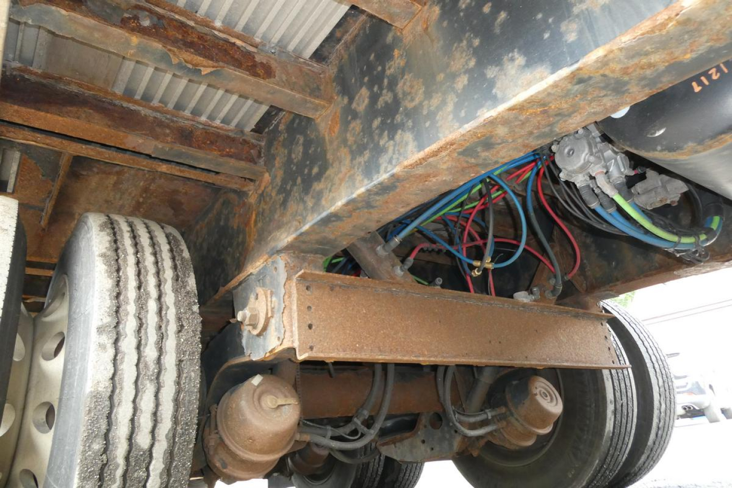 Underbody of trailer showing dual rear axles with leaf springs and wiring harness; rust-colored steel frame, air lines, and tire tread visible.