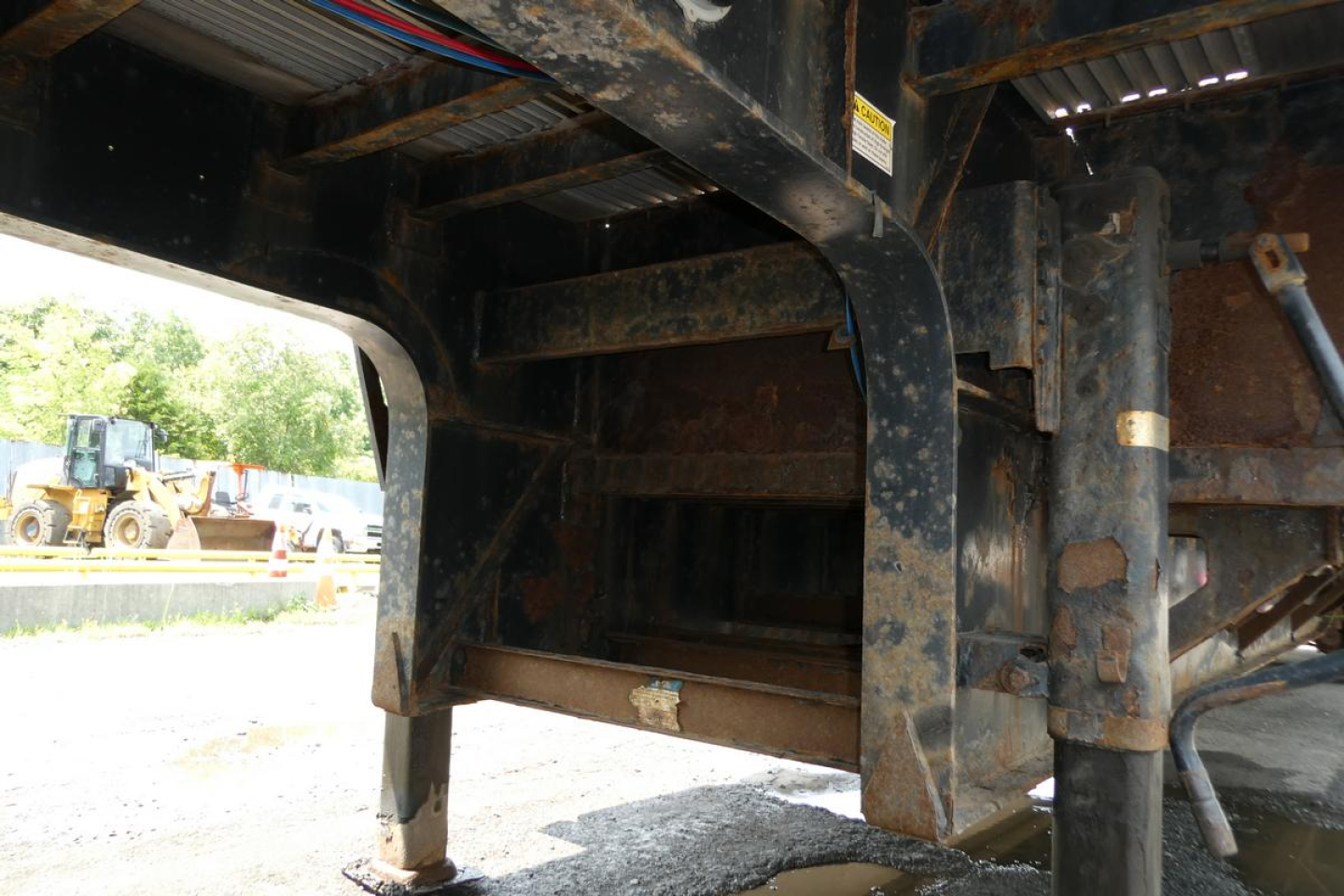 Underframe of a Fontaine HCITX22RSA drop deck trailer showing rusted steel frame and landing gear, with a storage shelf and hydraulic hoses nearby.