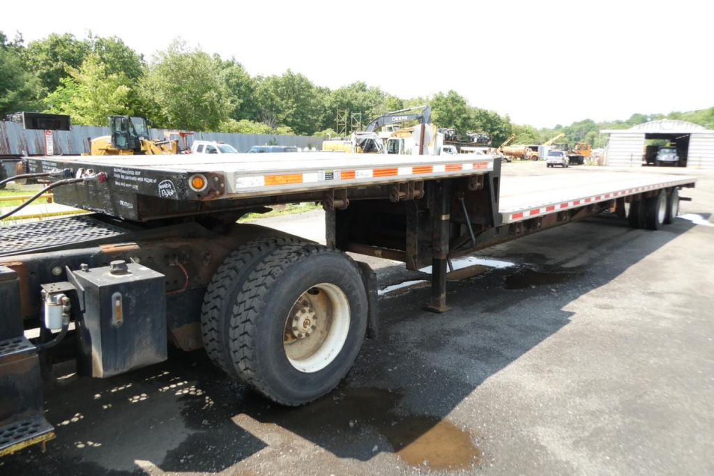 Flatbed trailer side view showing rear wheels and deck with reflective tape and hydraulic components.