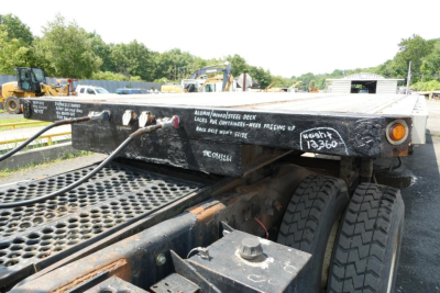 Trailer chassis with chalked text and hinges; visible on the deck: various handwritten notes, stock numbers, and a warning label. Dual rear tires and metal lattice deck visible.