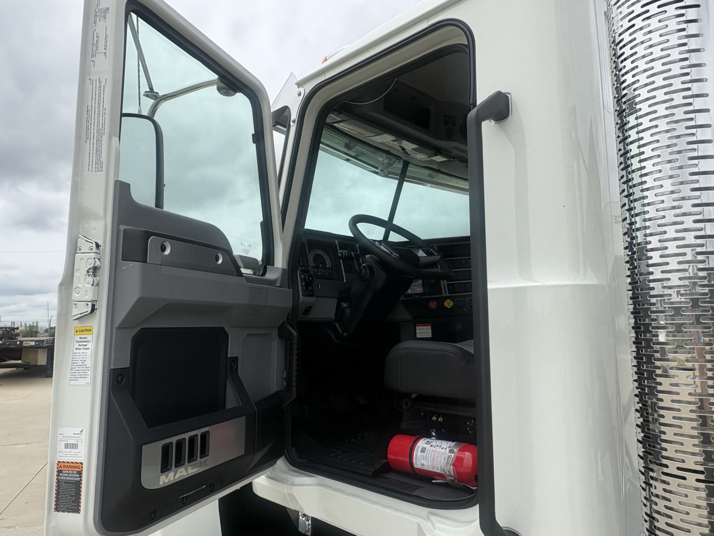 Truck cab interior with door open showing steering wheel, dashboard, and a red fire extinguisher on the floor; aluminum side panel with a woven metal door trim.
