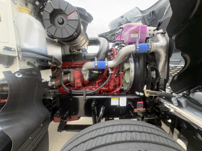 Mack PI64T engine bay showing red engine block, rubber hoses, coolant reservoir, and front axle with wheel; visible stickers and metal frame.