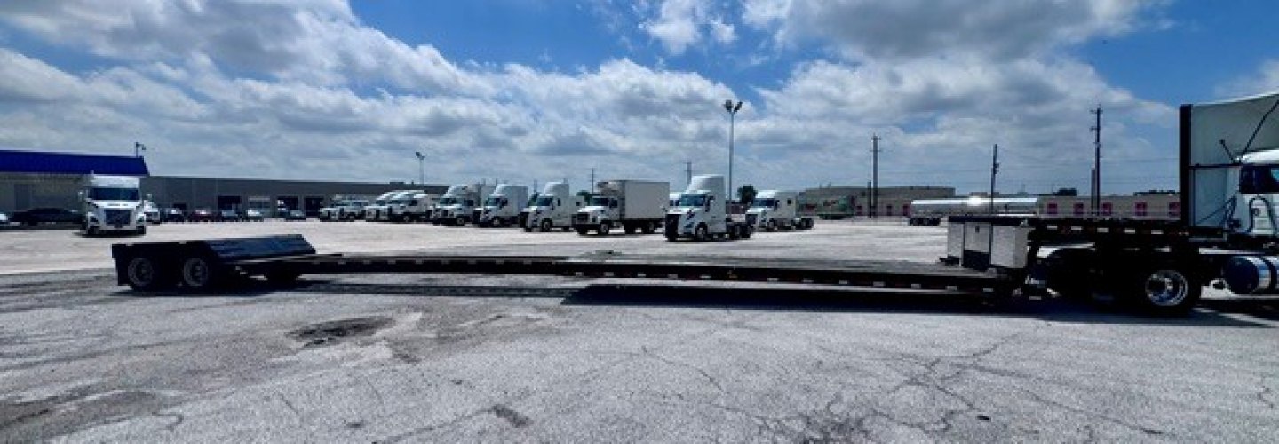 Lowboy trailer with long flatbed and truck tractors in background; visible signage and blue skies.