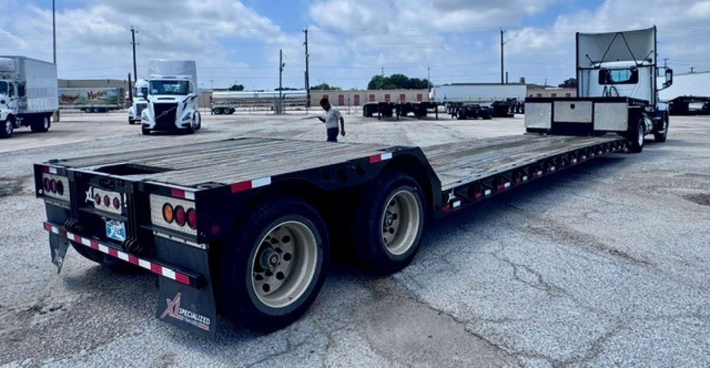 Lowboy trailer showing long flatbed with dual rear axles in a truck yard.