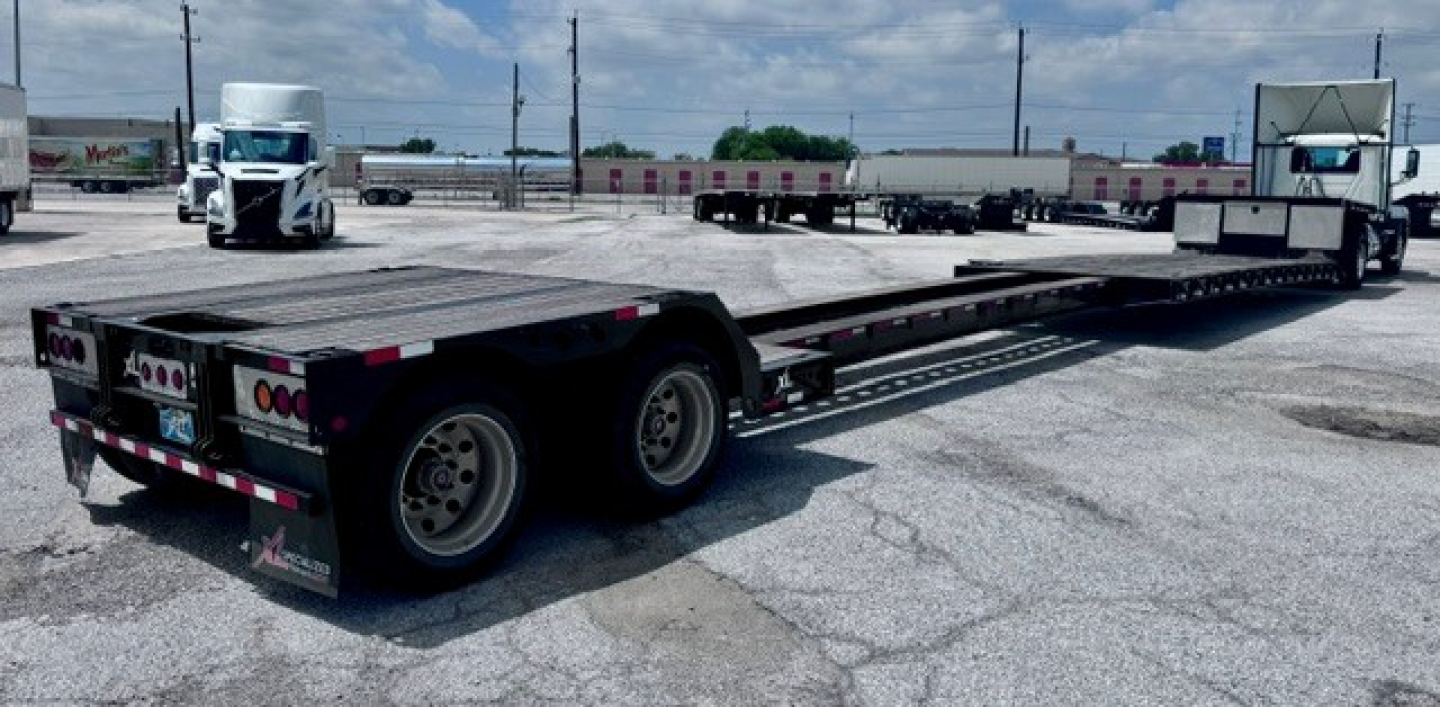 Lowboy trailer with extended flatbed and two rear axles visible, with semi-trailers and trucks in background; reflective safety tape on edges and landing gear visible.