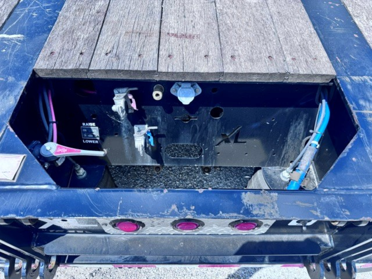 Trailer toolbox area with open access hatch showing battery cables and fittings, blue frame, metal panels, and wooden deck sections visible.