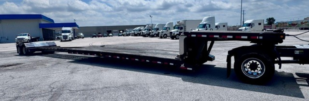 Lowboy trailer in a lot showing a long flatbed with a gooseneck hitch and heavy-duty suspension; clean surface with multiple axles and a wheel visible at the far end.
