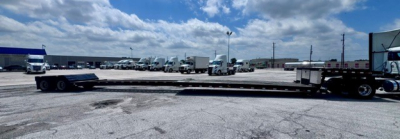 Lowboy trailer with long flatbed and truck tractors in background; visible signage and blue skies.