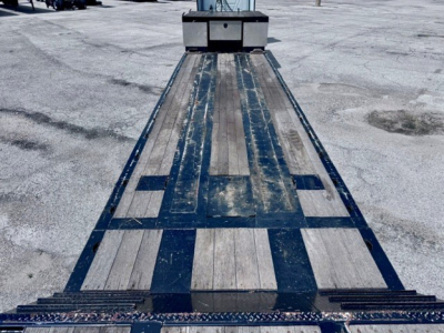 Flatbed trailer deck showing long wooden planks with metal crossmembers and loading ramps at rear, blue frame and weathered wood.