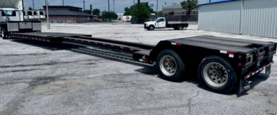 Flatbed lowboy trailer showing dual rear axles and long deck, black with diamond plate surface, rear ramps folded, in a gravel lot