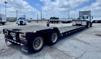 Lowboy trailer showing flatbed with multiple axles and rear tail lights; XL Specialized logo visible on mudflap; long equipment trailer in a lot.
