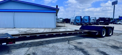 Lowboy trailer with flatbed deck and tandem axles in a lot.