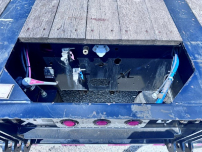 Trailer toolbox area with open access hatch showing battery cables and fittings, blue frame, metal panels, and wooden deck sections visible.
