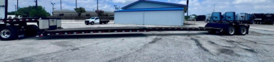 Long lowboy trailer with a blue revenue building in background. Side shows no legible text; general equipment view of a used trailer.