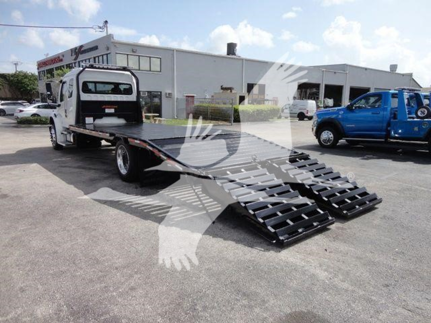 Flatbed rollback tow truck with extended dual ramps and Freightliner Business Class M2 cab in background.