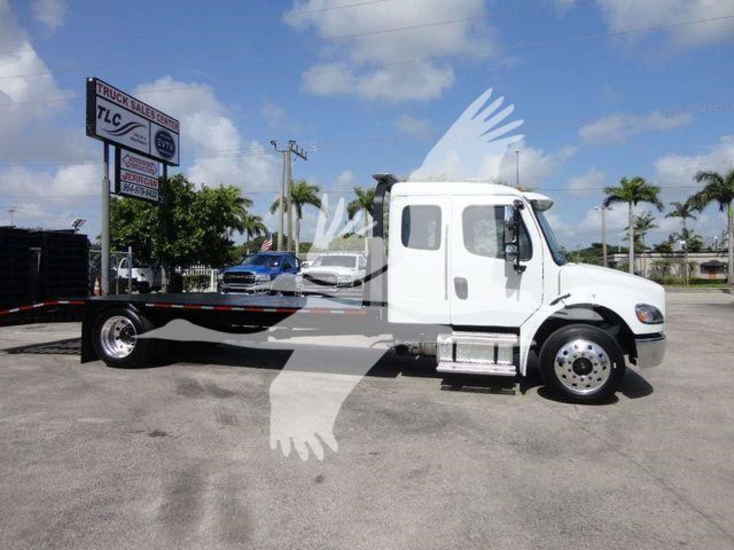Truck image: white Freightliner Business Class M2 rollback truck side view, cab with window, flatbed/dolly attachment, wheel rims, dealership lot background.