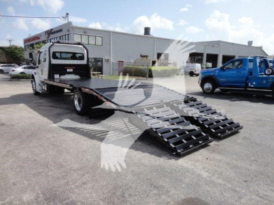 Flatbed rollback tow truck with extended dual ramps and Freightliner Business Class M2 cab in background.