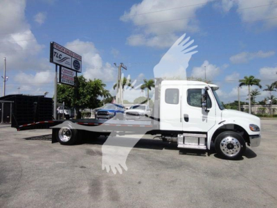 White Freightliner Business Class M2 rollback tow truck side view showing cab and flatbed with wheel, dealership sign in background