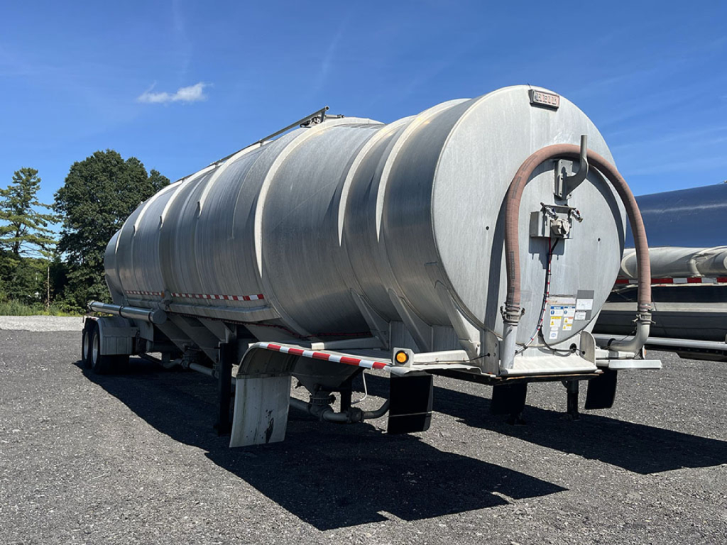 Aluminum pneumatic tanker trailer in-yard showing cylindrical tank with rear access hatch and hoses, front landing gear, and reflective tape.