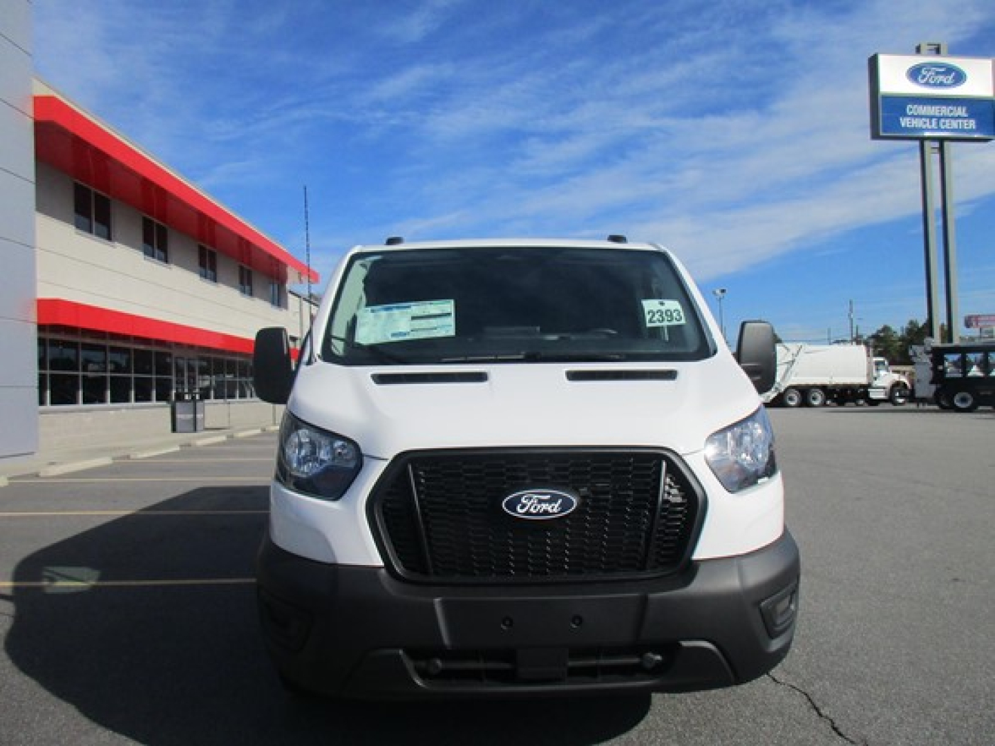 Front view of a white 2026 Ford Transit cargo van in a dealership lot with a large black grille and Ford emblem; windshield sticker visible.