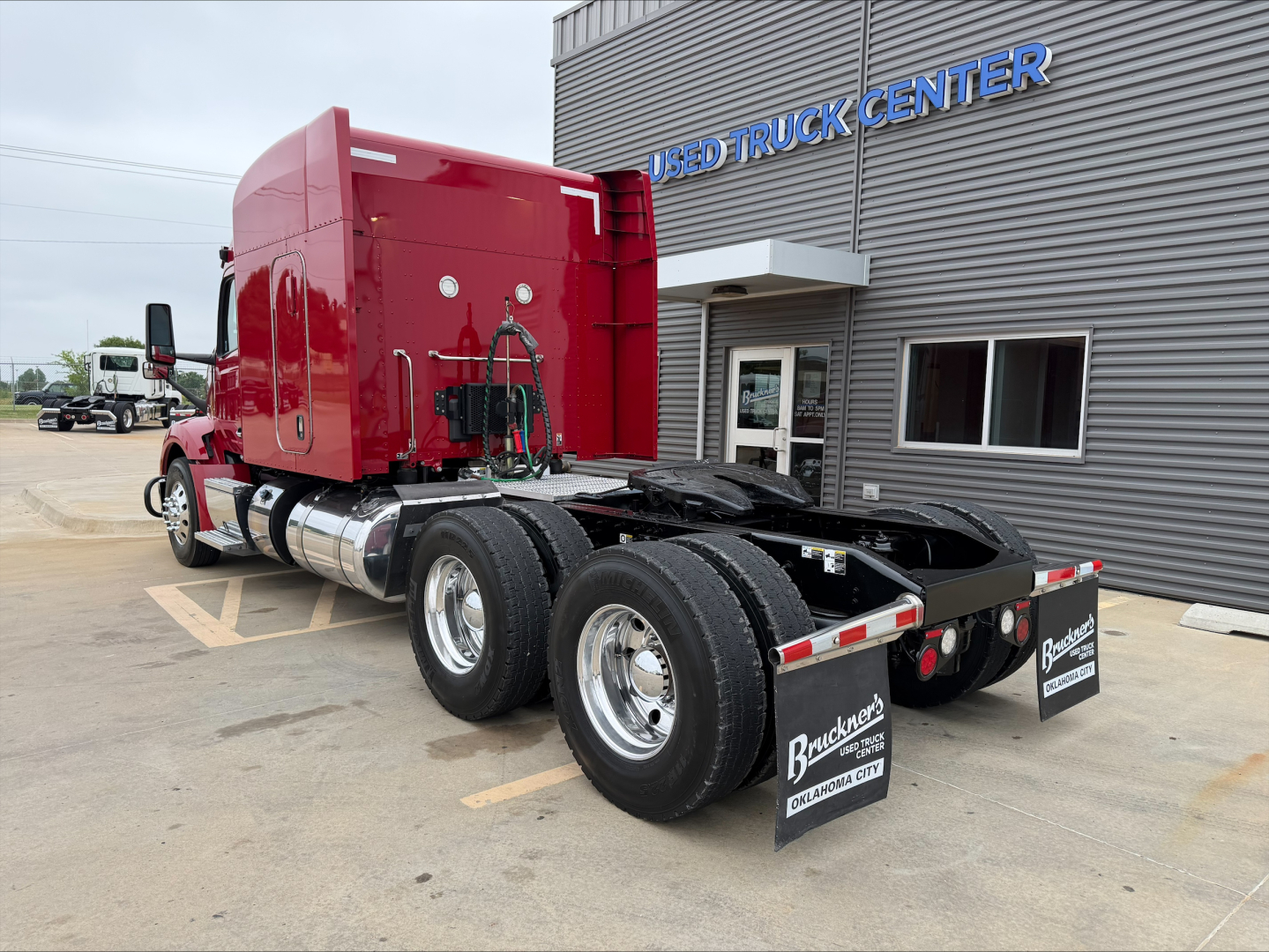 Peterbilt 579 tractor in red, rear view showing dual rear axles, fuel tanks, and mud flaps with ‘Backhus… Used Truck Center’ sign on building.