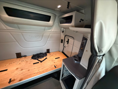 Peterbilt cab interior showing sleeper area with wooden floor panel, overhead storage, and control panel; Peterbilt logo on side wall.