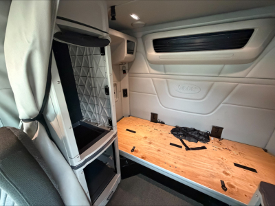 Peterbilt 579 truck interior with sleeper cab: empty side cabinet, plywood deck in cargo area, white padded walls, overhead light, and window vent panel.