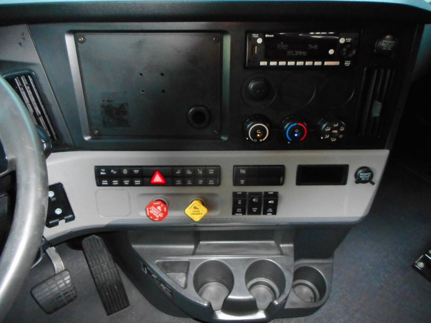 Truck dashboard and center console in a Freightliner Cascadia cab with steering wheel visible on left, various controls, cup holders, and a blank panel.